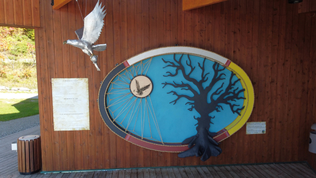On the exterior of a building, a circular art piece with white, yellow, red and black on the border of the circle. In the center a blue background, black tree and an image of a bird connected with strings. Hanging near the art piece is a silver bird.