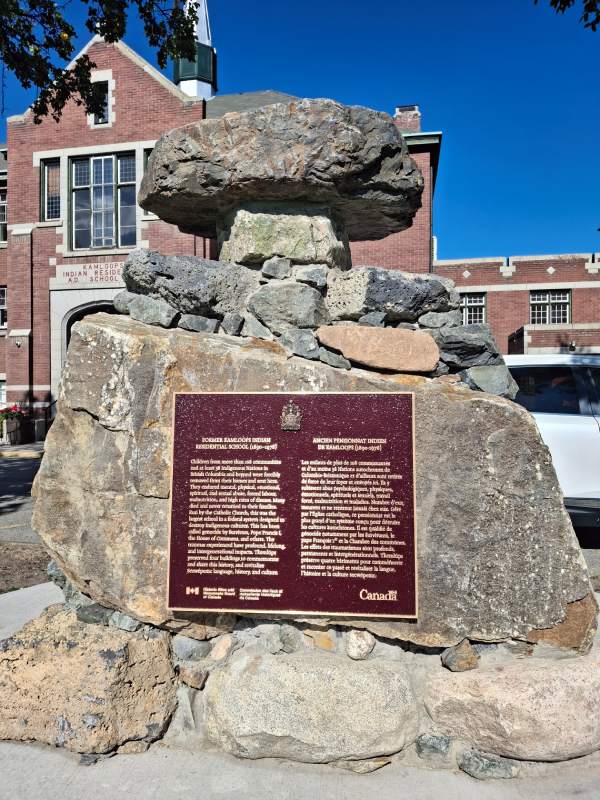 A commemorative plaque for the Former Kamloops Indian Residential School is installed on a monument made of stones, with a building in the background