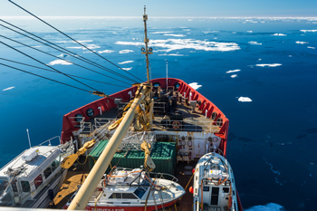 Front deck of icebreaker seen from above, sailing in blue sea with distant ice floes. Three smaller vessels sit on deck awaiting deployment.