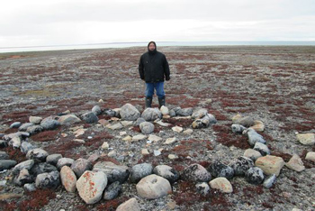 An Inuit man stands near a circular gathering of stones. The land around him is flat and barren.