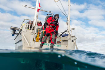 Diver suited in coldwater gear stands at the stern of a boat; half the image is underwater.