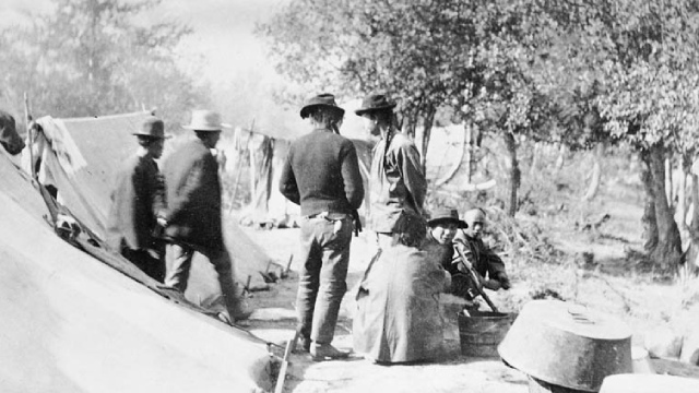 A black and white vintage photo of several people in a camp dressed in Victorian-era clothing.