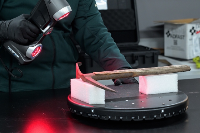 A person scans a historic axe with a handheld scanner in a lab, projecting red light on the object