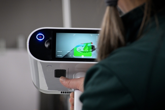 A person uses a handheld scanner to scan an artifact on a detailed calibration board, with the 3D model appearing on screen highlighted in green