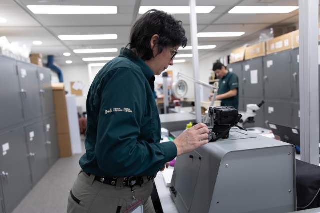 An archaeologist photographs an artifact with a specialized device, in a room where other specialists are working