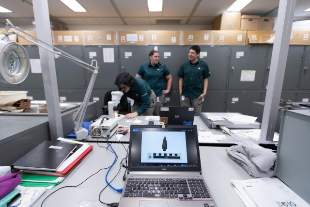 A projectile point is displayed on the screen of a laptop while archaeologists stand in the background