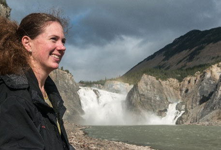 A smiling visitor stands on the shore of the Nahʔą  Dehé / South Nahanni River, with Virginia Falls behind them.