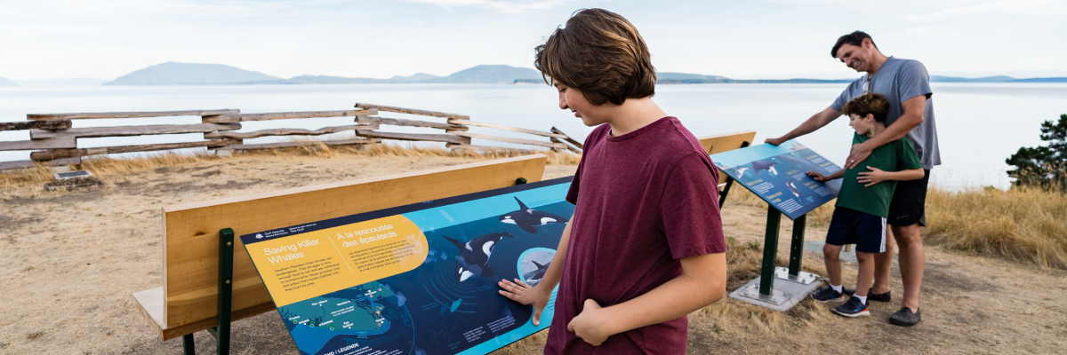 Un enfant, son père et son frère regardent deux panneaux d’interprétation à propos des baleines, avec la mer des Salish en arrière-plan.