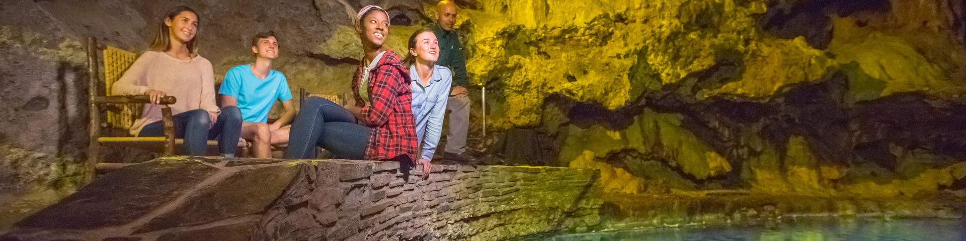 Des visiteurs assis aux abords des eaux thermales turquoise sont enchantés par l'expérience sensorielle qui leur est offerte, pendant qu'un interprète de Parcs Canada partage des histoires au sujet de la grotte avec ceux-ci. Lieu historique national Cave and Basin.