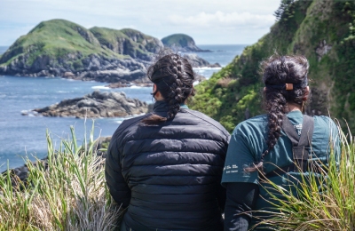 Deux femmes sont assises au bord d’une falaise qui surplombe une vue côtière et des îles.