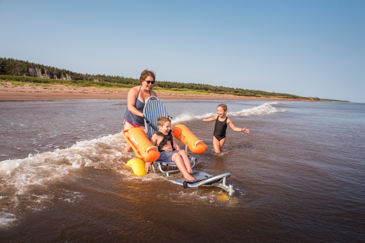 Une mère joue avec ses deux enfants dans les vagues à la plage. Un enfant est attaché à un dispositif d’assistance qui lui permet d’aller dans l’eau. 