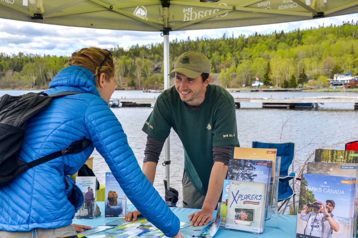 Un membre du personnel de Parcs Canada sourit sous un kiosque près de l’eau alors qu’il parle avec un visiteur.