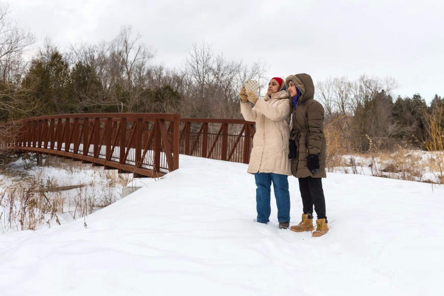 Deux personnes emmitouflées pour l’hiver s’arrêtent dans la neige pour prendre un égoportrait près d’un pont en acier et en bois.