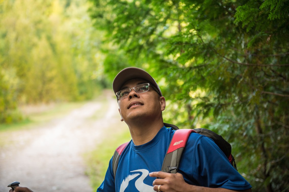 Gros plan d’une personne souriant, debout, tout en regardant vers le haut sur un sentier boisé.