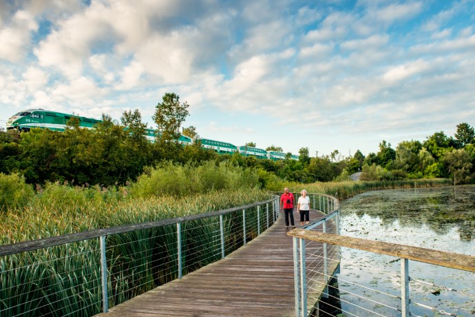 Two senior people walk on an enclosed wooden boardwalk over water, while a transit train passes in the distance.