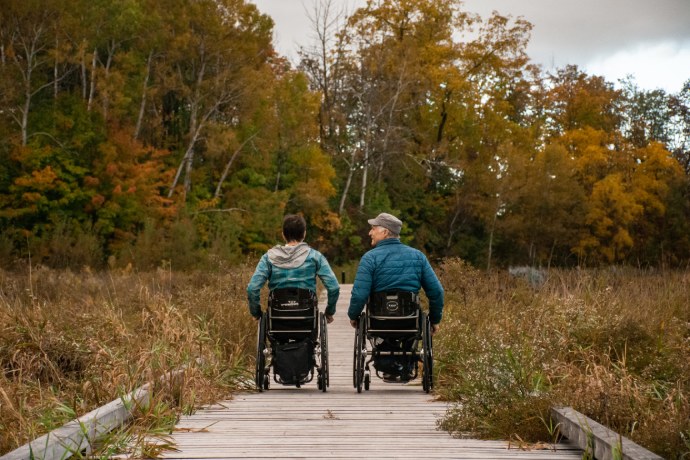 Two people in wheelchairs on a wooden boardwalk enjoy the fall forest and surrounding wetlands.