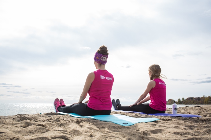 Two people sit on yoga mats on the beach as they look off toward the water.