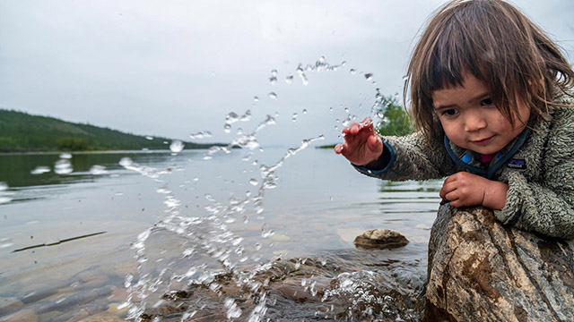 A toddler leaning over a rock by a lake with their hand splashing water droplets in the air.