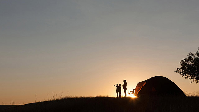Two children and an adult standing in front of a tent silhouetted by the setting sun behind them.