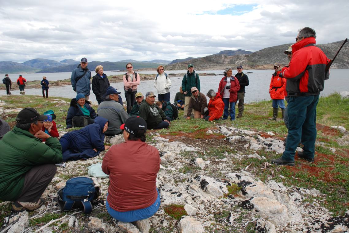 Un groupe de personnes assises ou debout près d’un plan d’eau entouré de montagnes devant un interlocuteur.