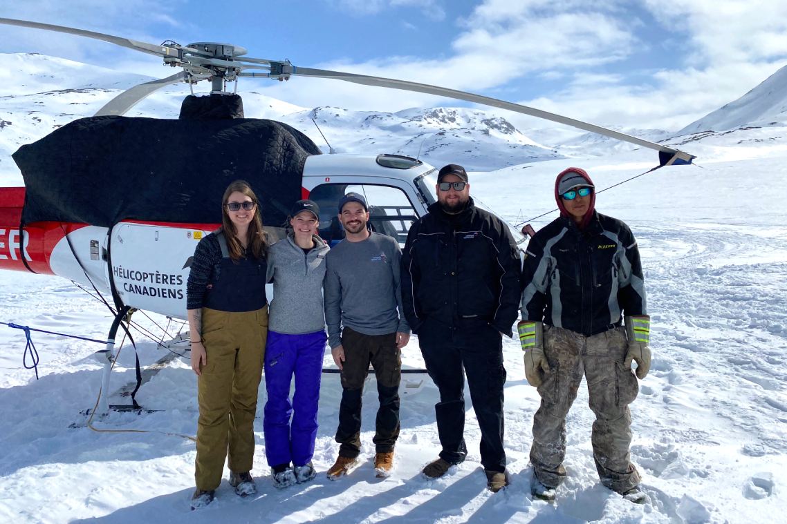 Five people stand together in front of a helicopter on the group in a wintry, mountainous landscape.
