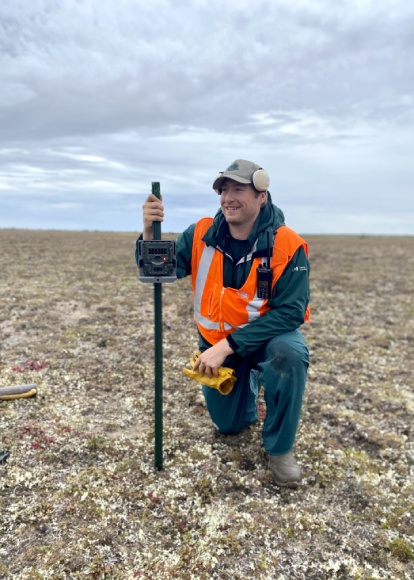 A Parks Canada employee wearing a safety vest kneels on one knee holding up a rod with a trail camera attached on a flat landscape.