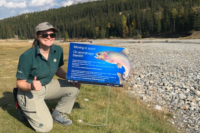 A Parks Canada staff person kneels down to give a thumbs up beside a small sign that reads Moving in soon.