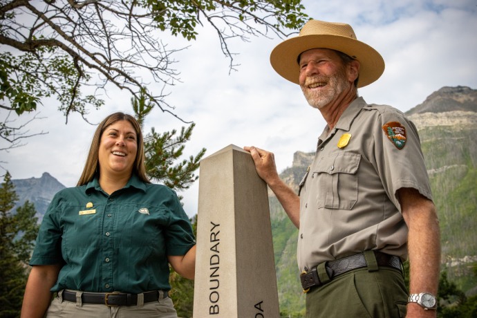 Un membre du personnel de Parcs Canada et du National Park Service des États-Unis se tiennent ensemble, chacun posant sa main sur un monument marqué Boundary (frontière). 