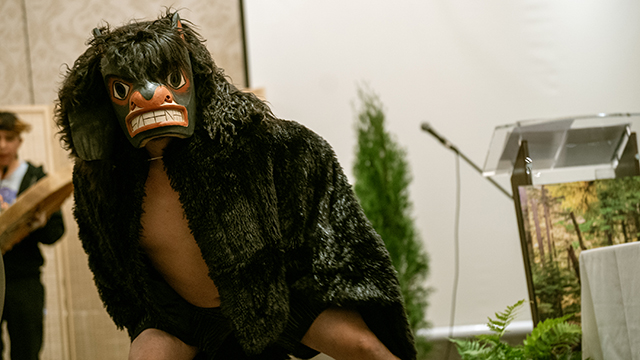 A person wearing a carved wooden mask and costume looks at the camera.