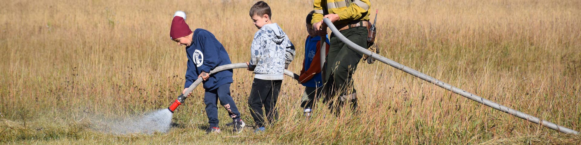 A fire crew member helps three children hold a fire hose as they spray down the grass. 