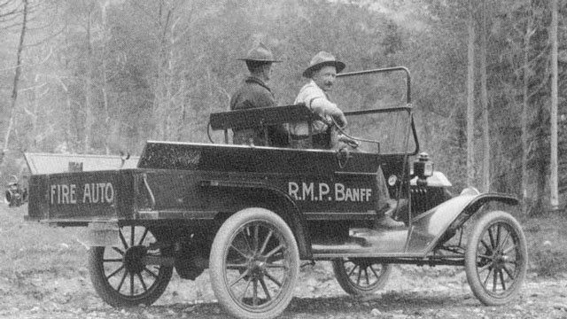 A historic image of Fire and Game Wardens in Banff National Park is shown in black and white. The men are sitting in an old vehicle with ‘Fire Auto’ written on the tailgate. 
