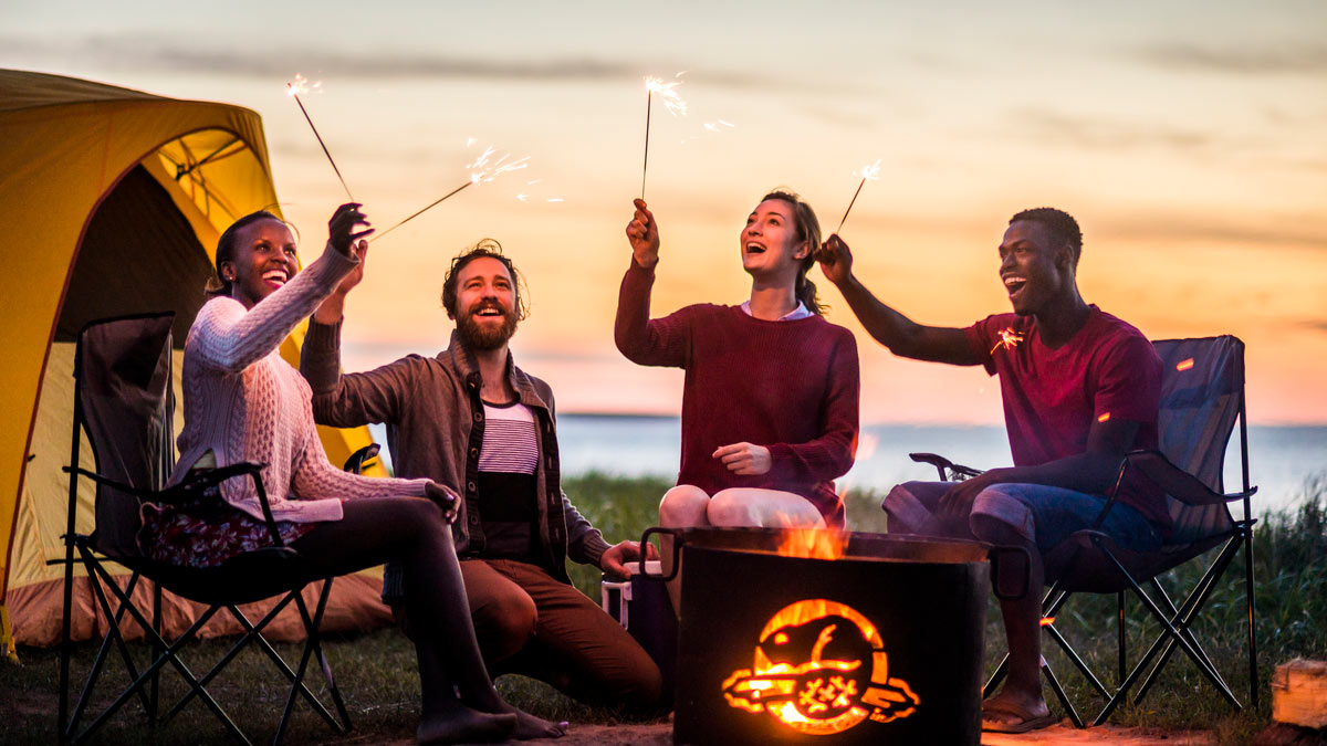 A group of friends light sparklers around a campfire during sunset at Cavendish Campground. Prince Edward Island National Park.