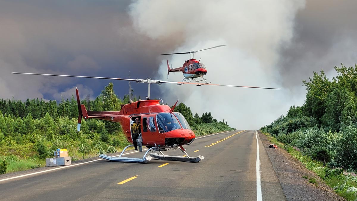 A helicopter sits on a closed highway. There is dense wildfire smoke in the background with a second helicopter taking off into the air.