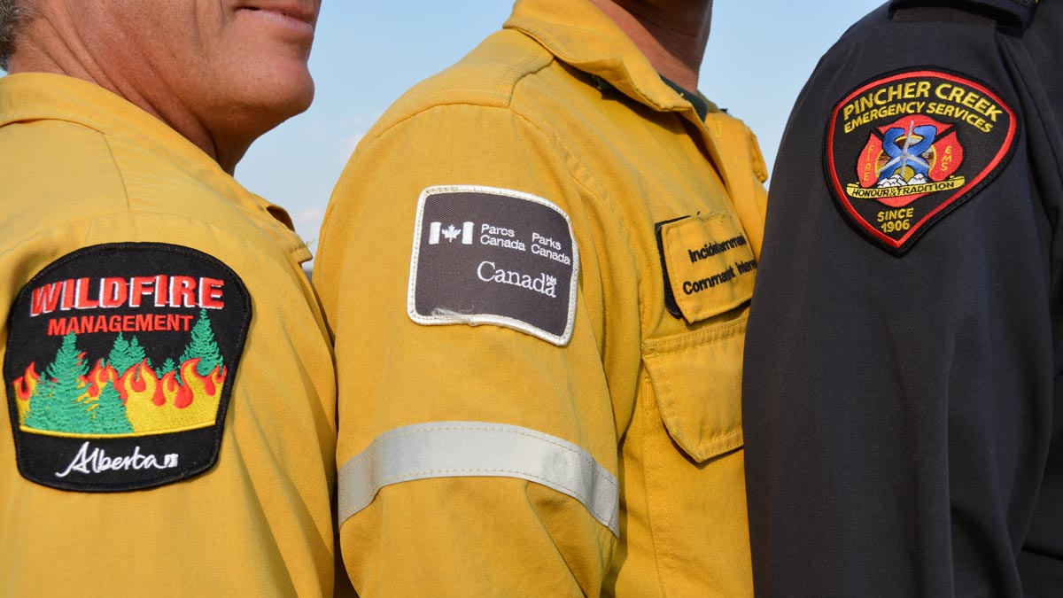 Three fire management specialists stand side-by-side during the Kenow wildfire in Waterton Lakes National Park. Shown are the arm patches from Alberta Wildfire, Parks Canada, and Pincher Creek Emergency Services.