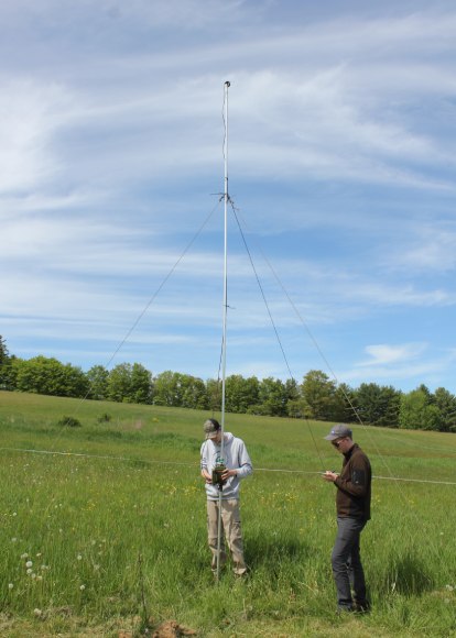 Two people record data next to a pole that extends high into the air in a grassy field.
