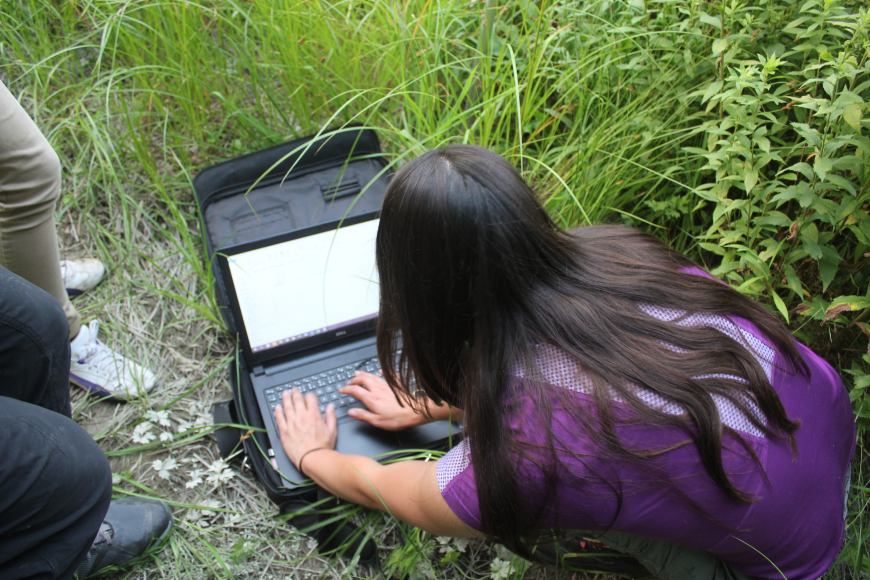 A close up of the back of a person typing on a laptop placed in a case on the ground outdoors.