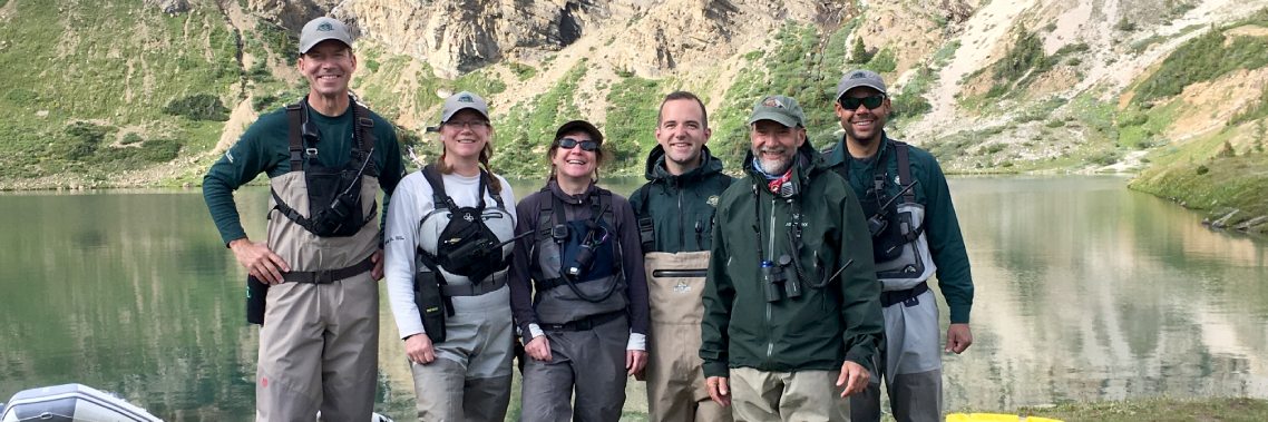 A group of six people dressed in outdoor work attire smile at the camera in front of a lake and mountainside.