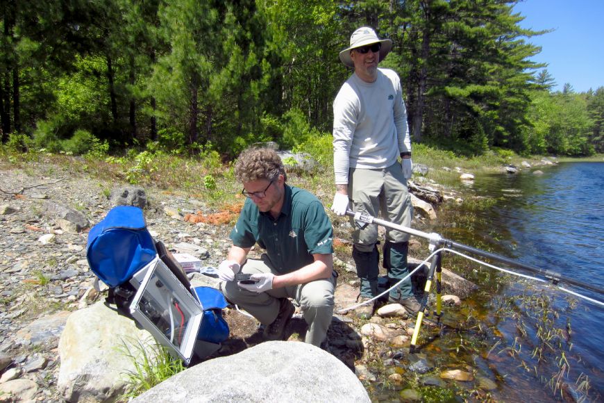 Two Parks Canada staff work at the edge of a lake. One is holding a supported beam into the water. The other is kneeling down wearing gloves while holding a device.
