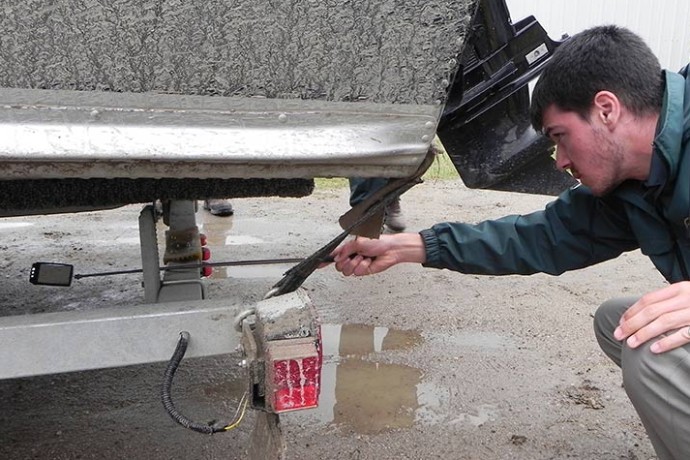 A Parks Canada staff member kneels down while extending an inspection tool beneath a boat trailer.