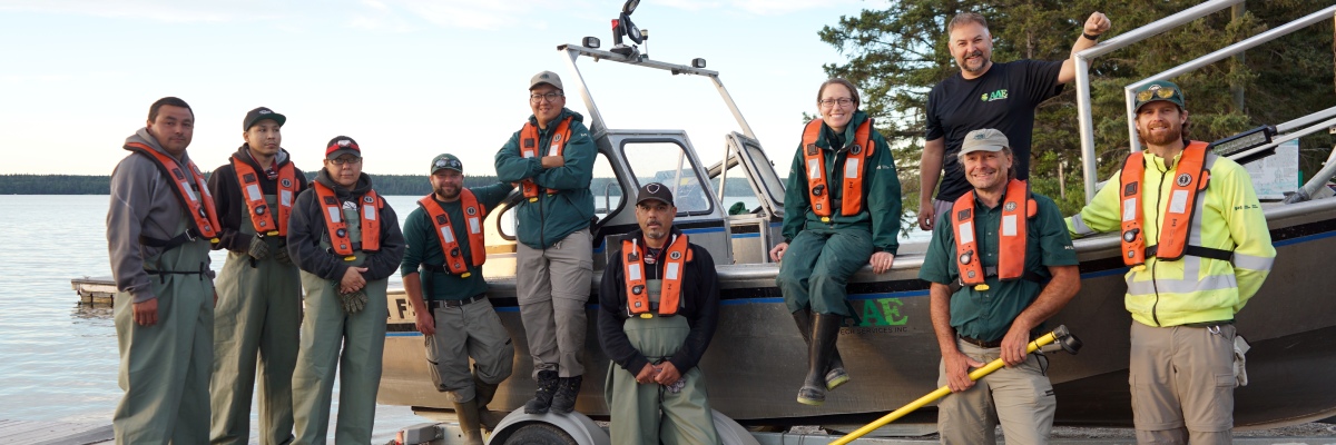 Un groupe de 10 membres du personnel de Parcs Canada et membres de la communauté autochtone sourient sur le rivage devant leur bateau de pêche électrique, qui est mis à l’eau sur une remorque.