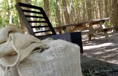 Un sac en toile de jute rempli de bois est placé à côté d'un foyer en métal dans un terrain de camping.