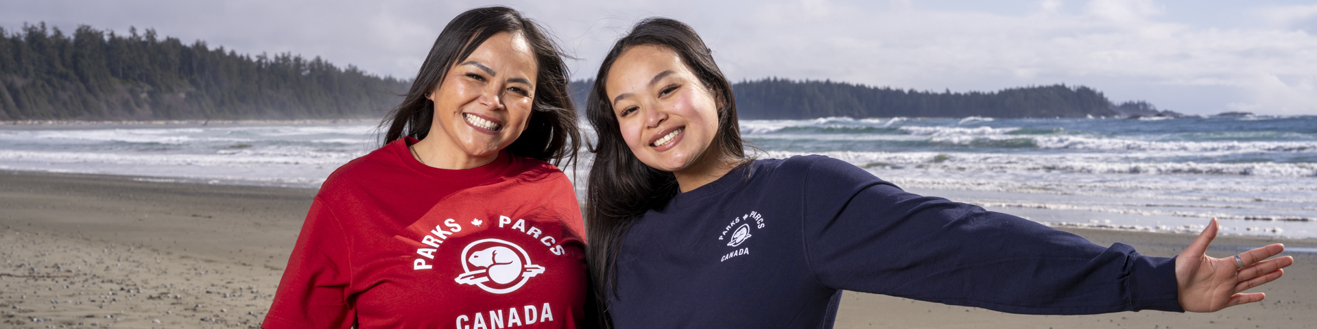 Two women smiling and spreading their arm are wearing Parks Canada official merchandise while on a beach with the ocean in the background.