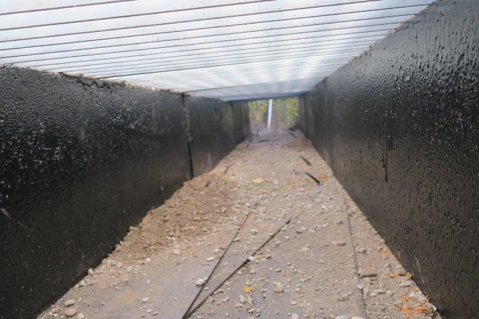 Une vue de l’intérieur du tunnel souterrain montre un sol en terre battue, des murs en ciment et un plafond en grilles métalliques.