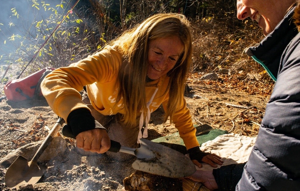 A person cooks over a campfire with a shovel while another assists. They are outdoors in a wooded area.