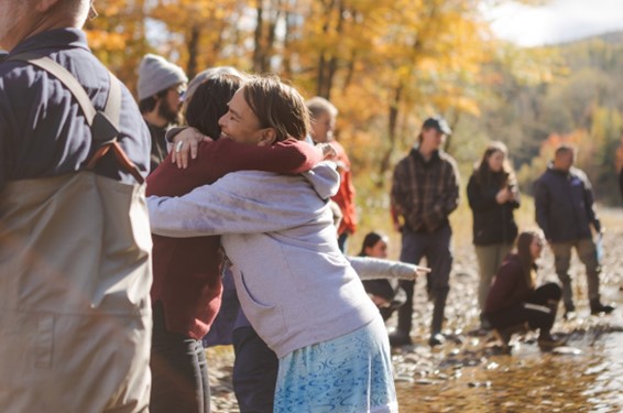 People hug by a riverbank as others stand and sit nearby.