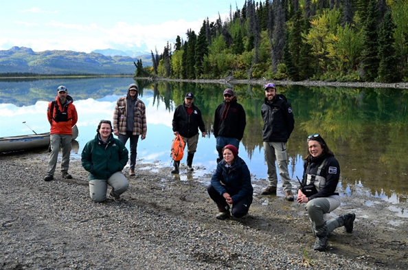 A group of people in outdoor gear gather by a calm lakeside with forested hills in the background.