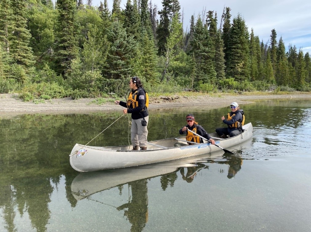 Three people in life vests are in a canoe on a calm river surrounded by dense trees. One stands while the others are seated.