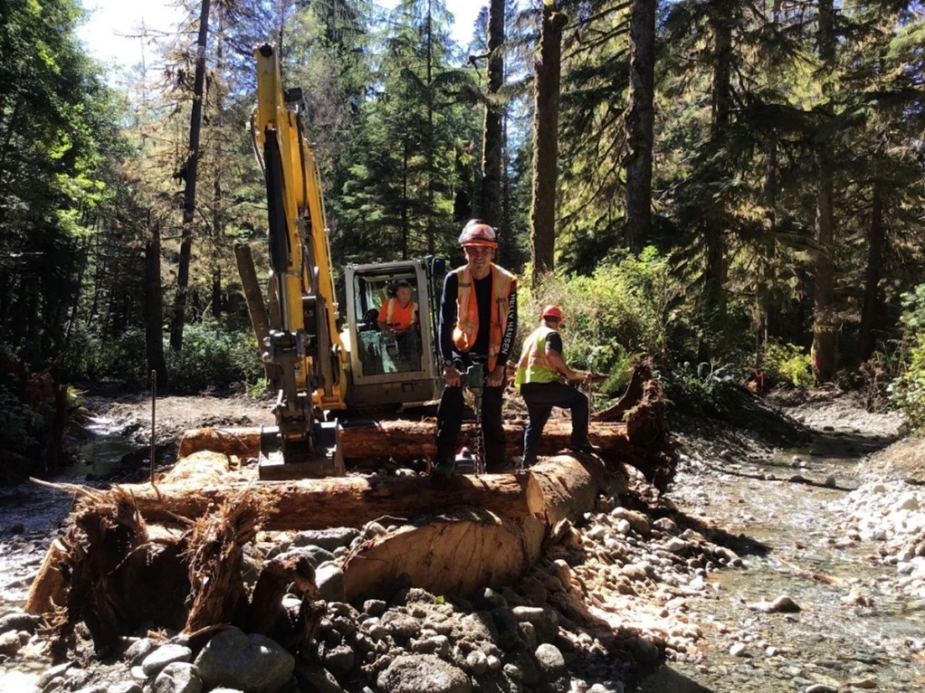 Workers in safety gear operate an excavator on logs in a forested area by a rocky stream.