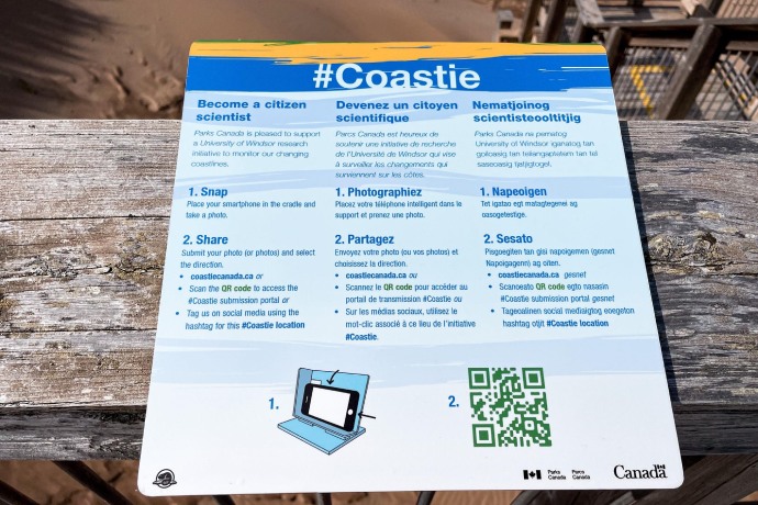 A close up view of a sign attached to a wooden boardwalk on a sandy beach, giving instructions in English, French, and Mi'kmaq on how to take a Coastie.