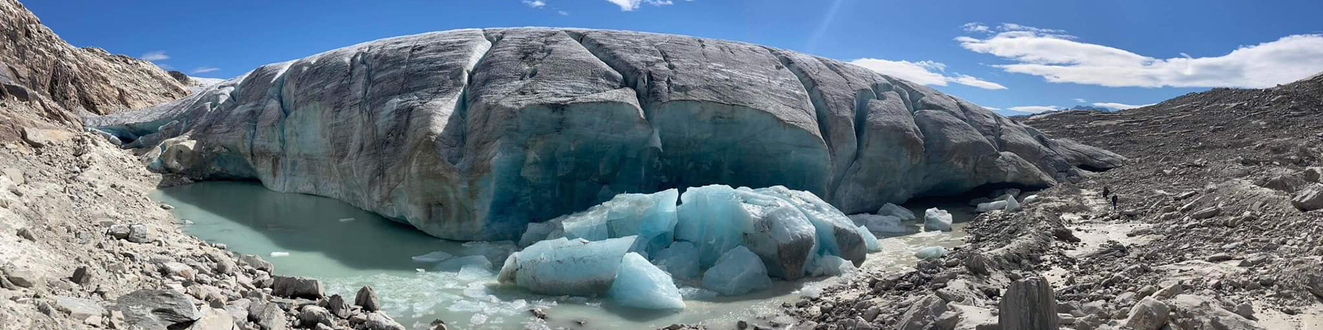 A panoramic view of Hilda Glacier in a rocky mountainous landscape. A glacial meltwater stream with a silty texture winds through the barren valley. Snow-covered peaks and rugged terrain dominate the background under a clear blue sky.